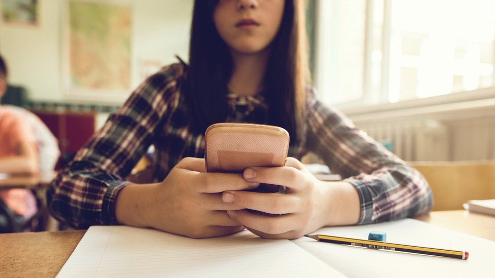 Stock image of a close up of schoolgirl typing text message on a mobile phone at a desk in aclassroom