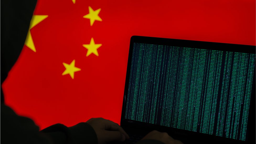 Stock image of hooded person working on computer in front of a Chinese flag