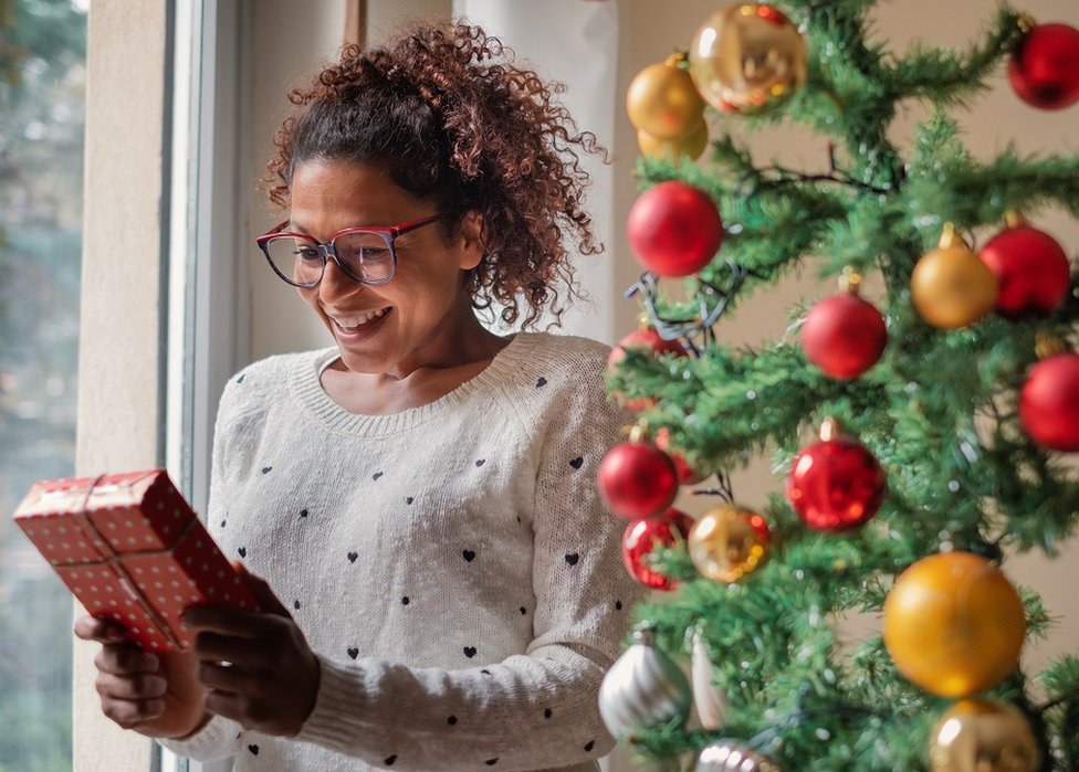 Happy woman standing next to a window and a Christmas tree, excited before opening her gift