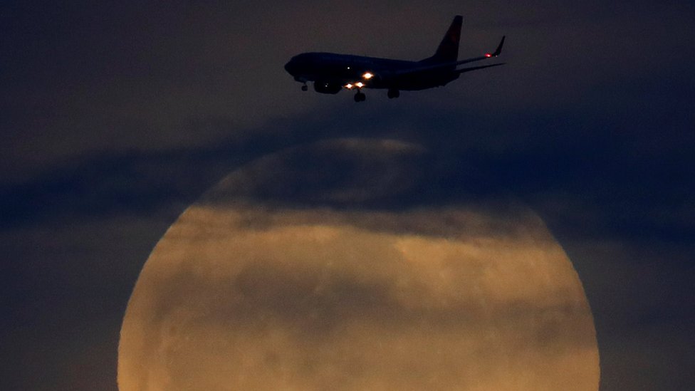 A full moon rises between clouds as a landing commercial jet approaches the airport before the start of a total lunar eclipse that is called a 