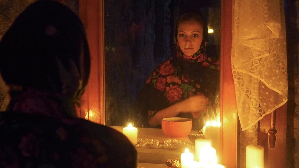 A girl standing in front of a mirror, it's dark and she's using candles for some light.