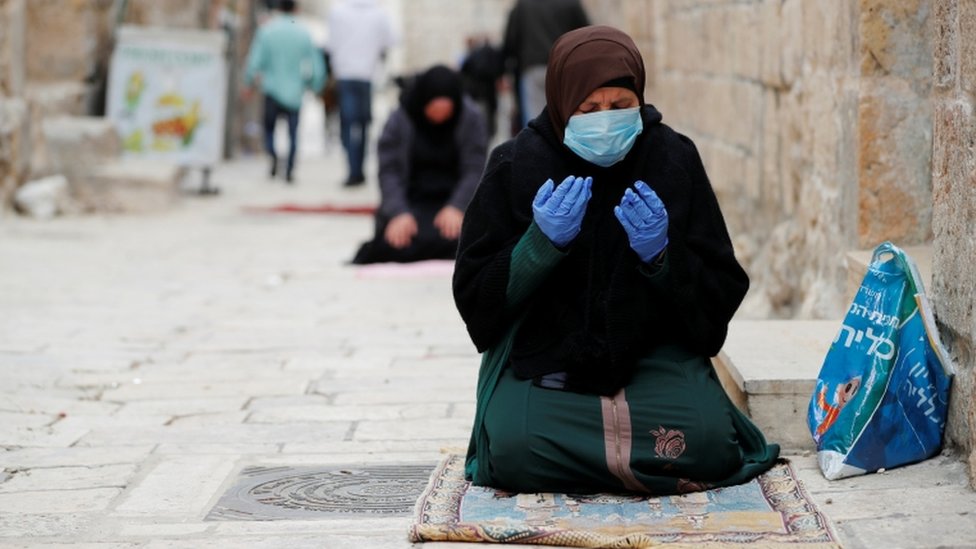 A Muslim woman wearing a face mask and rubber gloves prays
