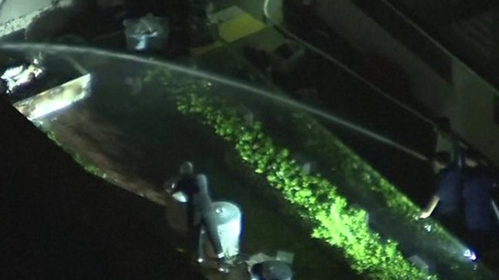 People spraying water on object outside of consulate and man closing rubbish bin