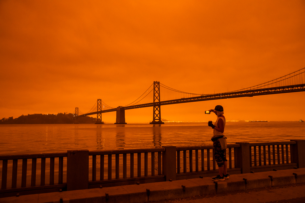 9 ستمبر 2020 کو سان فرانسسکو میں جنگلوں میں لگنے والی آگ کے بعد ایک خاتون اس کی تصویر لیتے ہوئے۔ آسمان مکمل طور پر نارنجی رنگ کا نظر آ رہا ہے۔