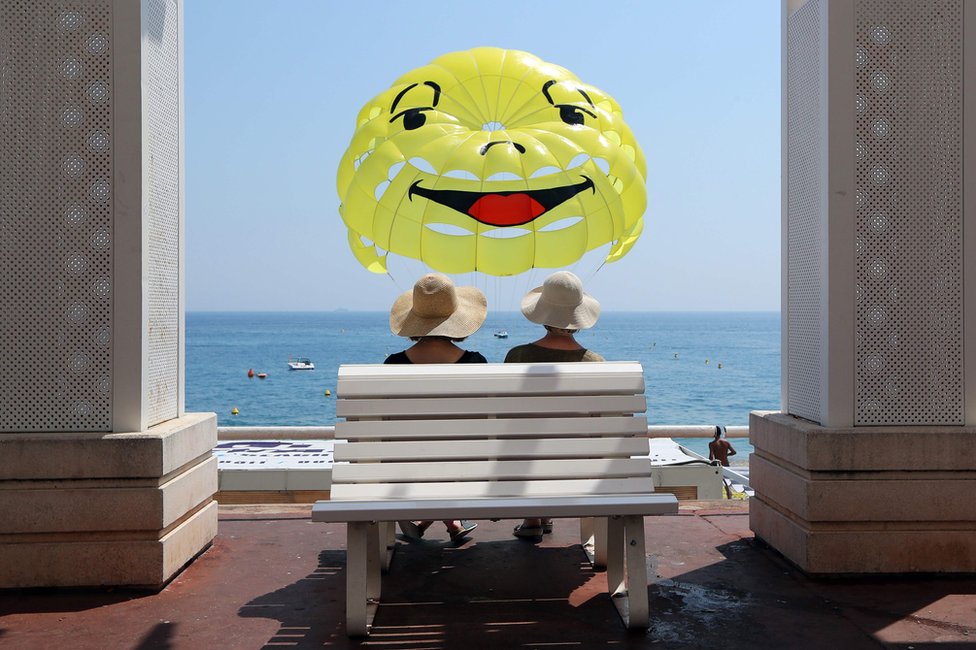 People sit on a bench while people parasail on the beach in Nice, on the French Riviera