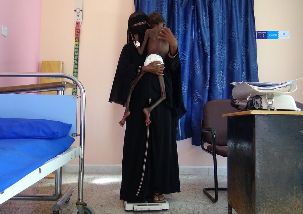 A woman holds her child whilst she stands on a scale