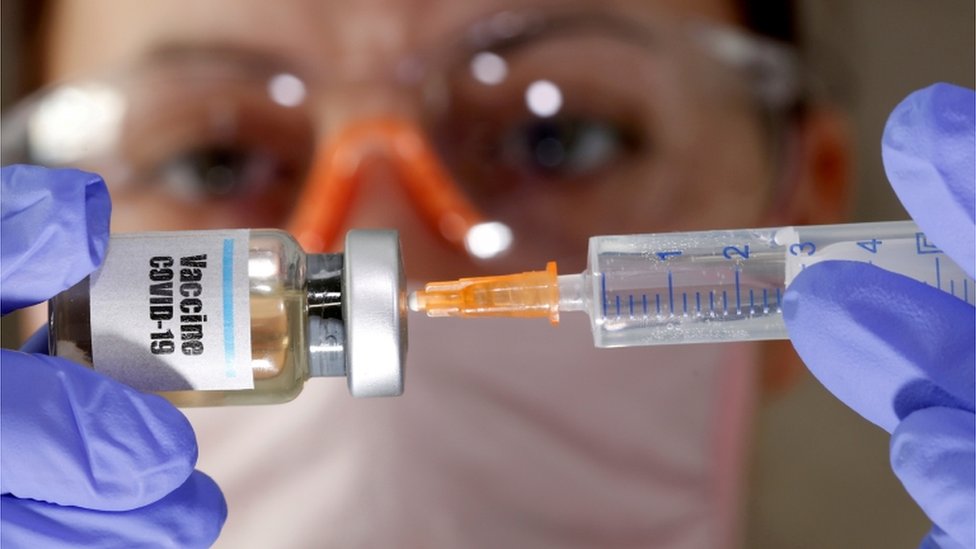 Photograph of a woman wearing gloves and PPE putting a syringe into a bottle marked Covid-19