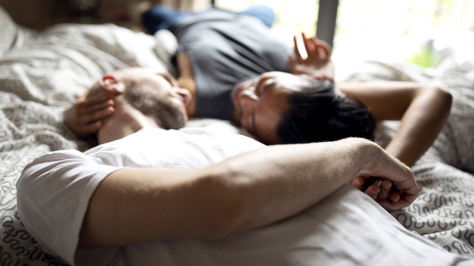 Two men gaze into each others' eyes lying on a bed