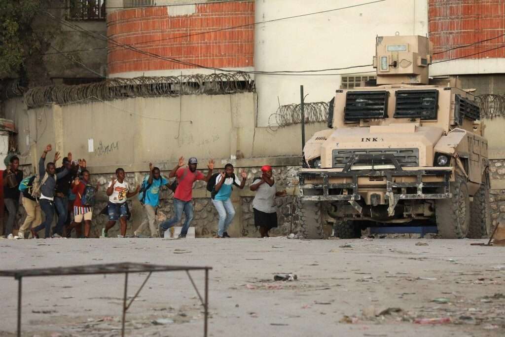 People walking for cover with their hands in the air towards a military vehicle